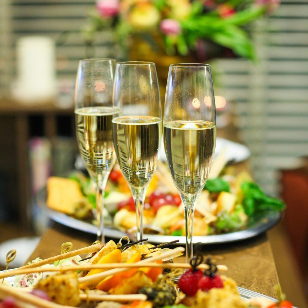 Trzy kieliszki z bąbelkami na tacy z przekąskami (serami, owocami) w tle, symbolizujące uroczystość.