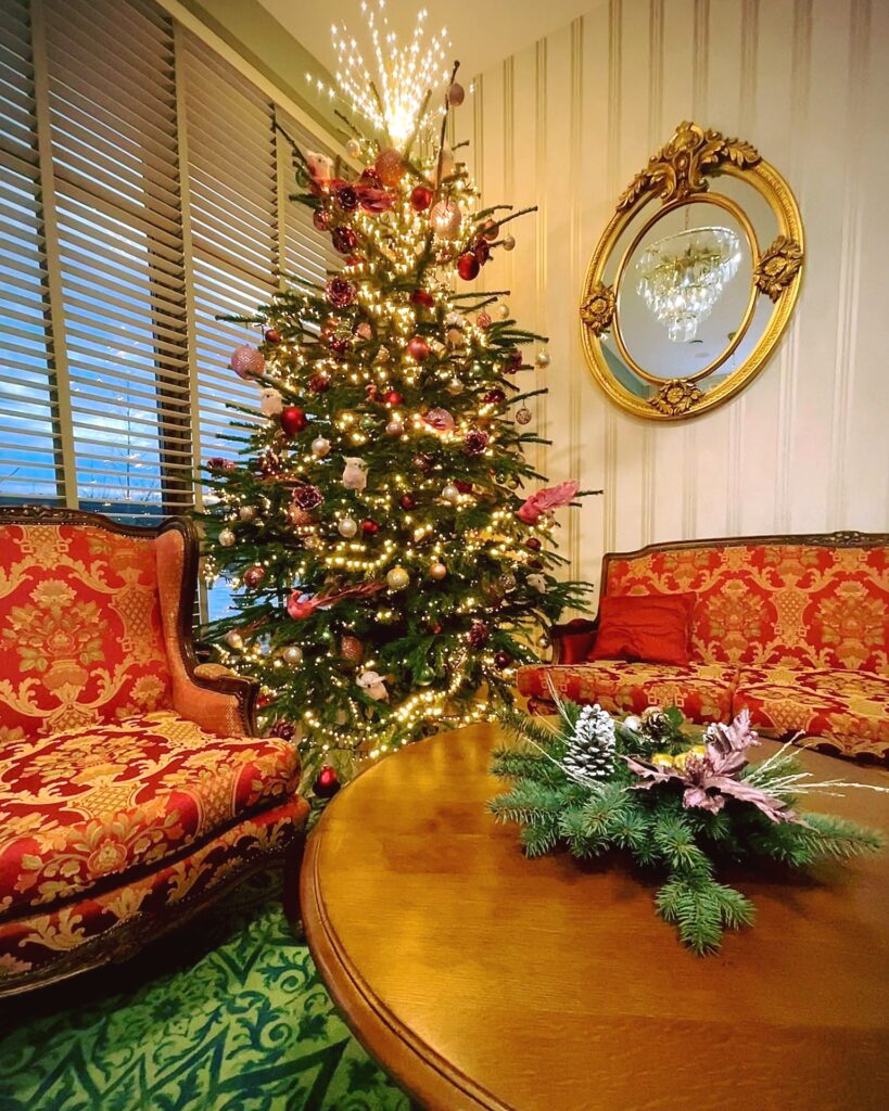 Wnętrze eleganckiego salonu z czerwoną sofą i fotelami, ozdobionego choinką bożonarodzeniową i lustrem w złotej ramie.