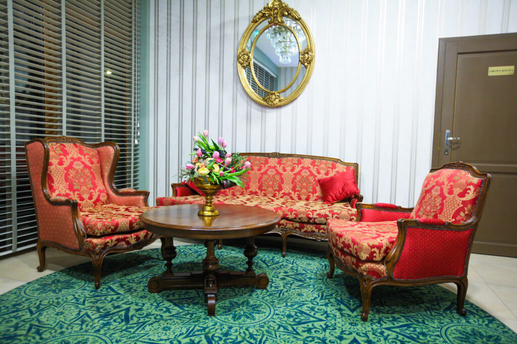 Wnętrze eleganckiego salonu z czerwoną sofą i fotelami, ozdobionego choinką bożonarodzeniową i lustrem w złotej ramie.