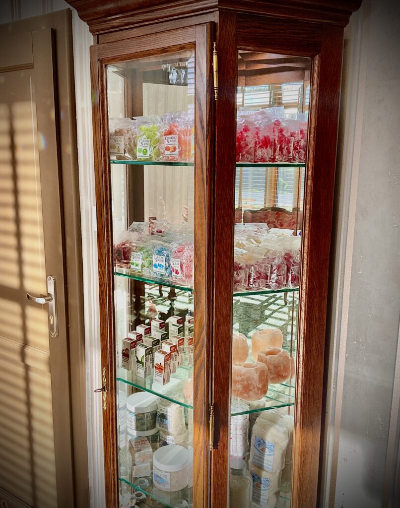 Drewniana gablota z przeszklonymi drzwiczkami, w której znajdują się różnorodne produkty solne, takie jak sole do kąpieli, kosmetyki i lampy solne