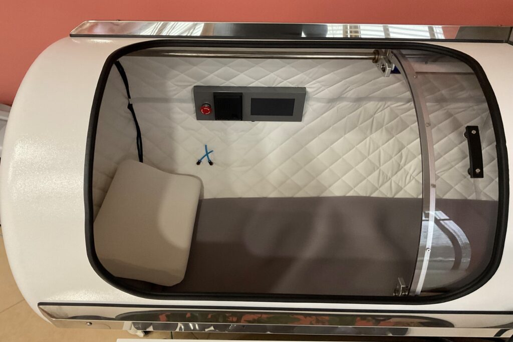 Wnętrze komory hiperbarycznej z białymi, pikowanymi ściankami, szarym materacem, panelem sterowania i małą poduszką